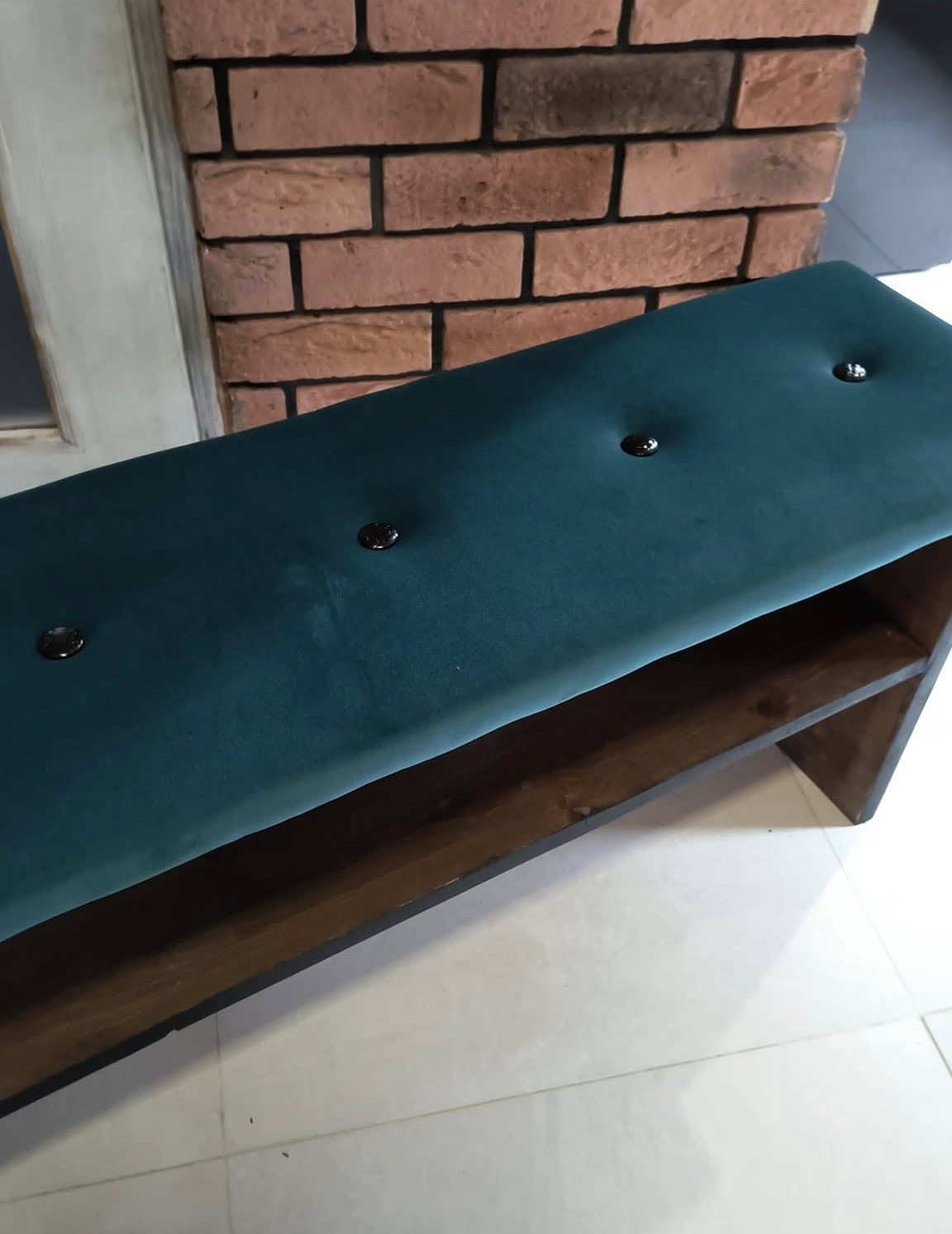 Tapicerowana ławka z ciemnozielonym welurem i drewnianą podstawą, wykończona guzikami, na tle ceglanej ściany.