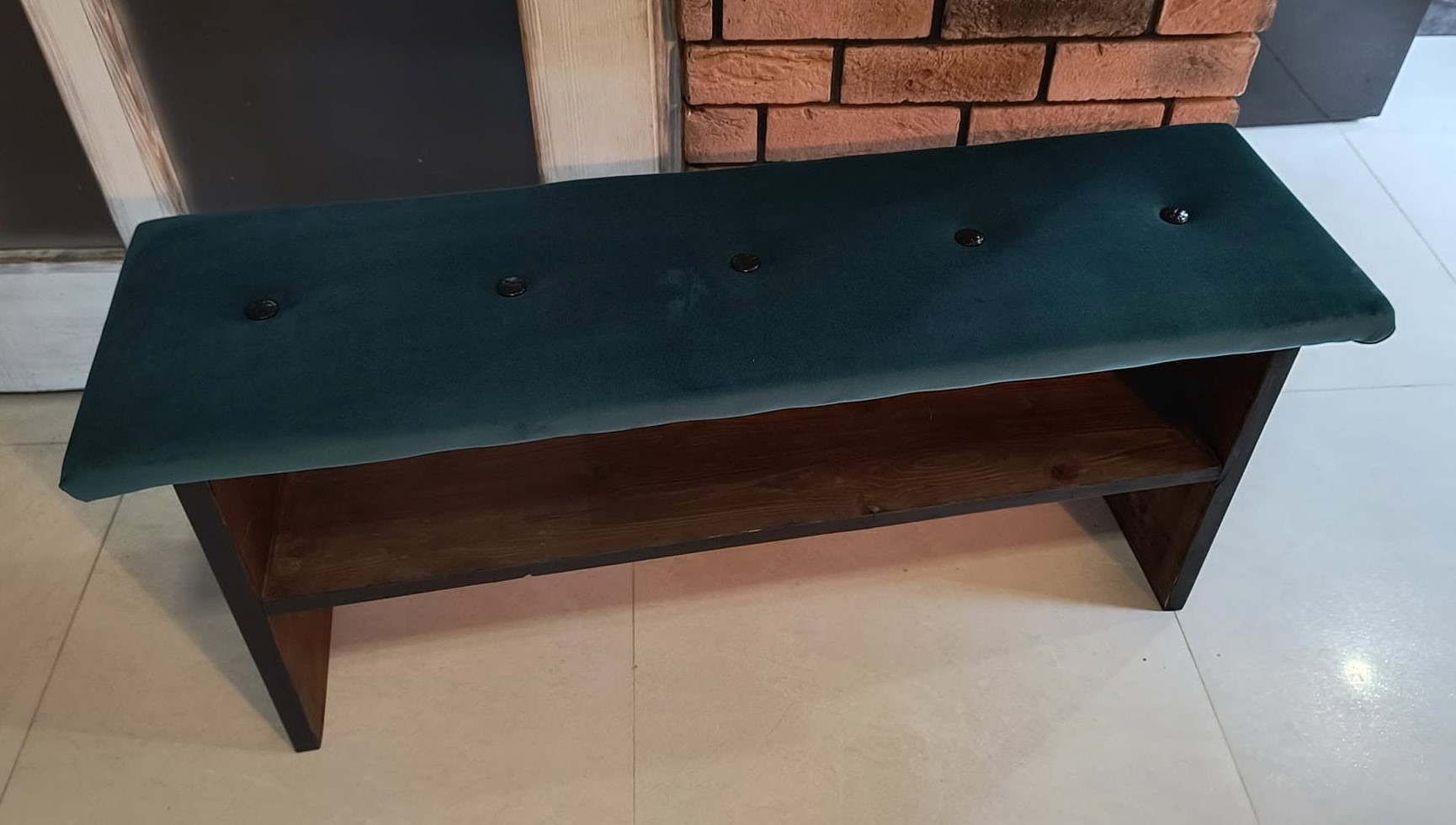 Tapicerowana pufa w kolorze butelkowej zieleni z drewnianą półką, wykończona guzikami, na tle ceglanej ściany.