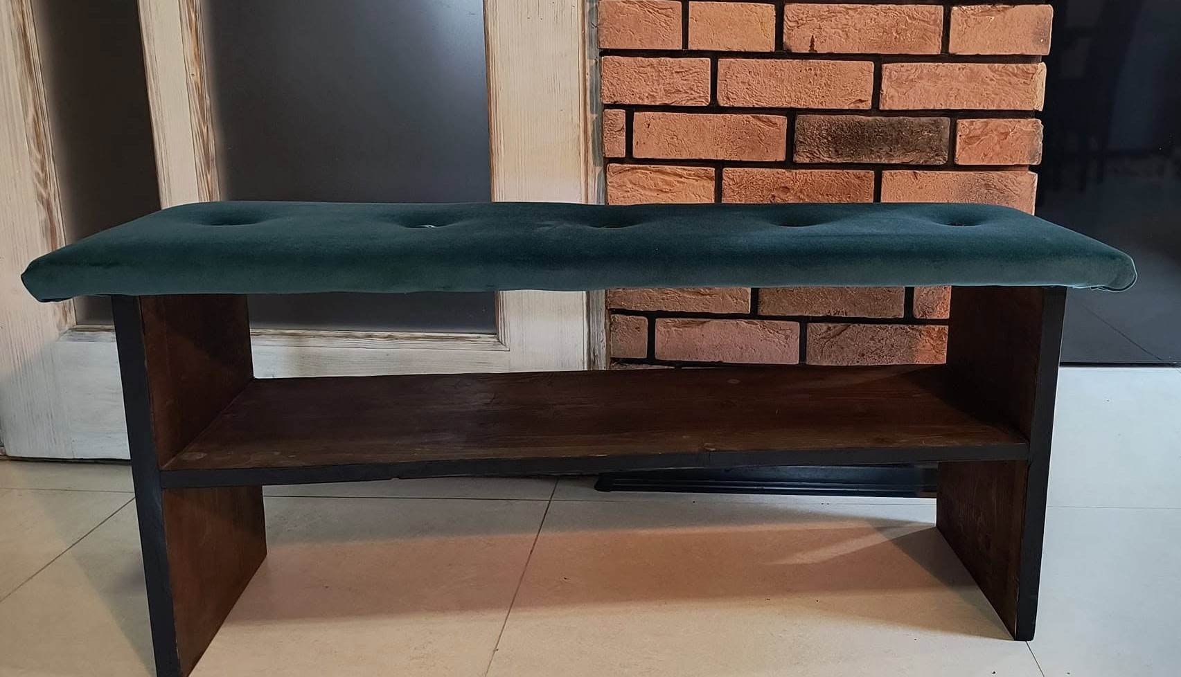 Tapicerowana pufa w kolorze butelkowej zieleni z guzikami, na drewnianej ramie z półką, na tle ściany z cegły i drzwi.