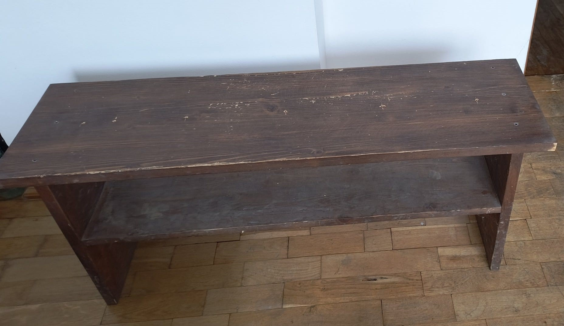 Stary, drewniany stolik kawowy z półką, widoczne ślady użytkowania i odpryski farby, ustawiony na parkiecie.