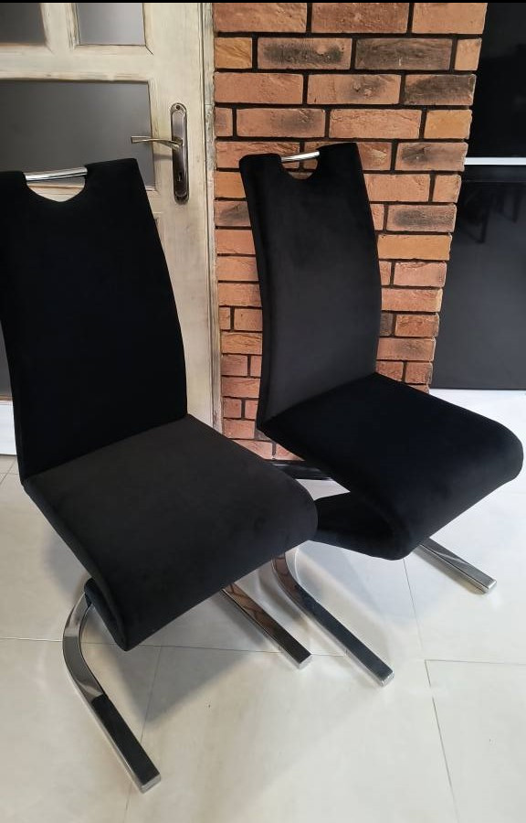 Dwa krzesła tapicerowane czarnym welurem z chromowanymi nogami, ustawione na białej podłodze przy ceglanej ścianie i białych drzwiach.