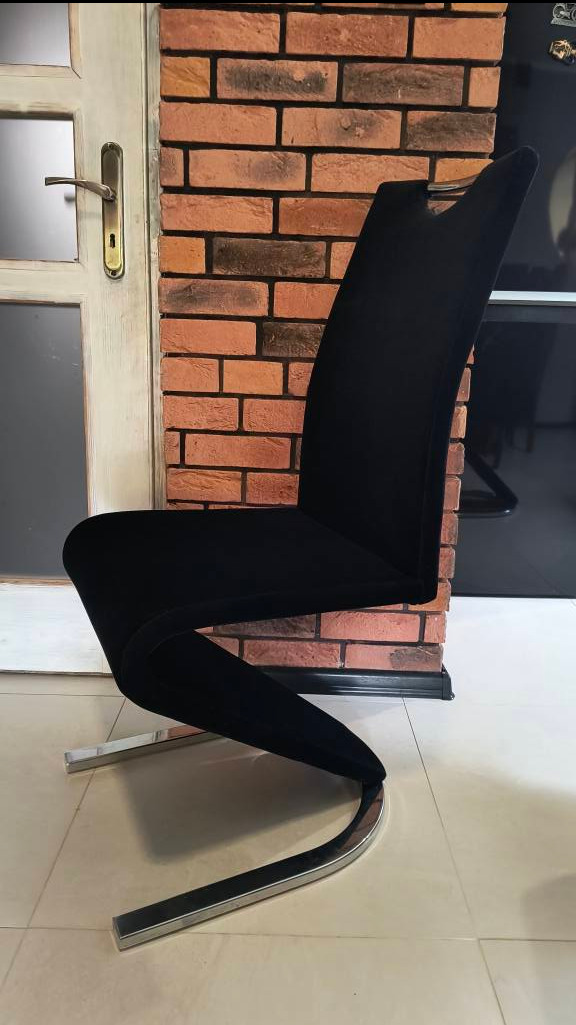 Eleganckie czarne krzesło z chromowaną podstawą w kształcie litery S, ustawione na jasnej podłodze na tle ceglanej ściany i białych drzwi.