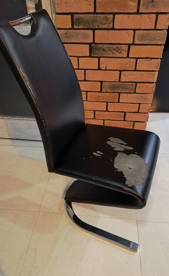 Uszkodzone czarne krzesło tapicerowane z chromowaną podstawą, widoczne ślady zużycia i odchodząca farba na siedzisku, na tle ściany z czerwonej cegły.