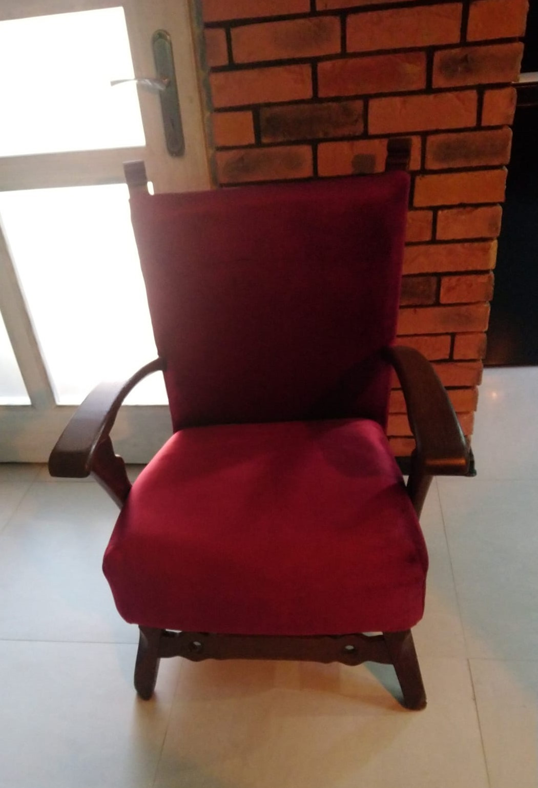 Odnowiony fotel z bordową tapicerką, drewniane podłokietniki, na tle ceglanej ściany i białych drzwi.