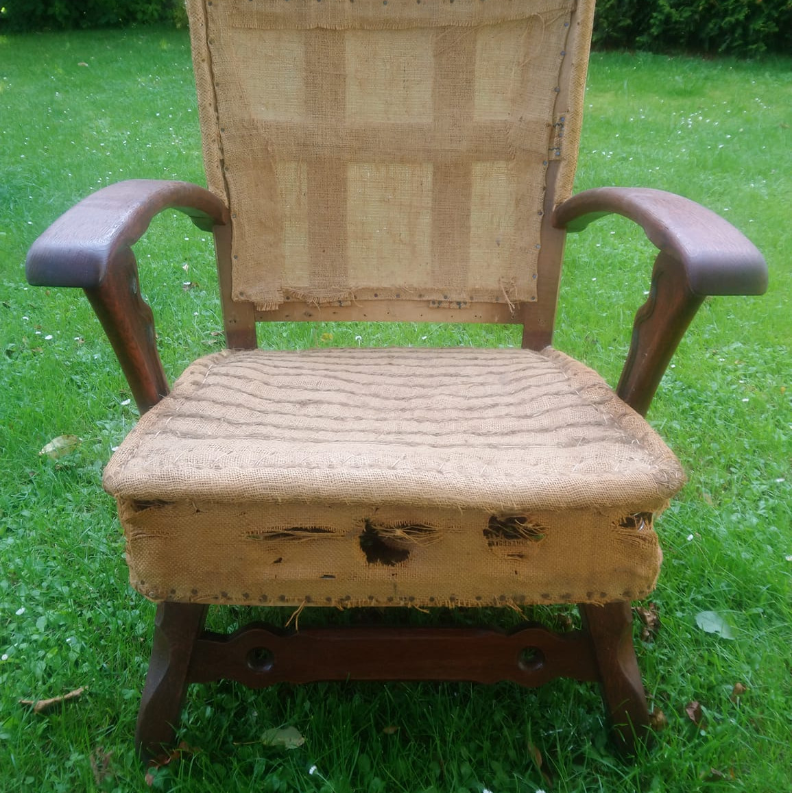 Stare, zniszczone krzesło z drewnianymi podłokietnikami i jutowym obiciem, ukazujące potrzebę renowacji, ustawione na trawie.