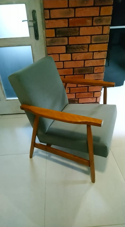 Odnowiony fotel w stylu vintage z drewnianymi podłokietnikami i szarozieloną tapicerką, ustawiony na białej podłodze z płytek, z fragmentem ściany z cegły dekoracyjnej w tle.