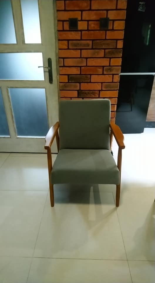Odnowiony fotel z szarą tapicerką i drewnianymi podłokietnikami stoi na jasnych kafelkach, na tle drzwi i ściany z cegły.