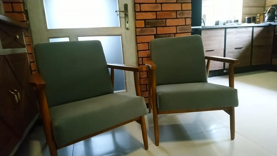 Dwa odnowione fotele w stylu retro z drewnianymi ramami i szarym obiciem, stojące na jasnej podłodze w Rabce-Zdroju, z widoczną ceglaną ścianą w tle.