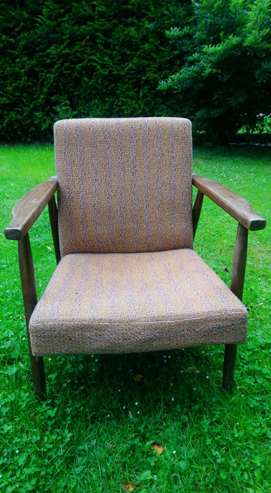 Fotel z drewnianymi podłokietnikami i tapicerką w odcieniach brązu, stojący na trawie w ogrodzie.