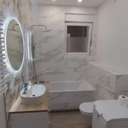 Nowoczesna łazienka po remoncie: marmurowe płytki, okrągłe lustro z podświetleniem LED, drewniany blat umywalki, prysznic za szklaną ścianką, pralka i toaleta.