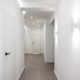 STRONG WALL SP. Z O.O. - Nowoczesny korytarz z szarą podłogą z płytek, białymi ścianami i drzwiami, oświetlony ledowymi lampami sufitowymi i kinkietami.