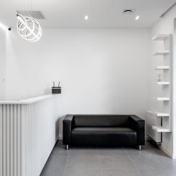 STRONG WALL SP. Z O.O. - Nowoczesne wnętrze: biała recepcja z dekoracyjnym oświetleniem, czarna skórzana sofa i minimalistyczne białe półki.