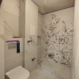 STRONG WALL SP. Z O.O. - Wnętrze łazienki w trakcie wykończenia, widoczne białe marmurowe płytki z motywem kwiatowym, podwieszana toaleta z zabezpieczeniem, wystające kable i rury, fioletowa taśma malarska na górze ścian.