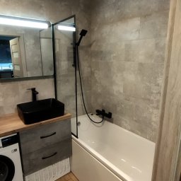 STRONG WALL SP. Z O.O. - Nowoczesna łazienka z beżowymi płytkami imitującymi kamień, czarną armaturą, prostokątną umywalką nablatową, parawanem nawannowym i drewnopodobną podłogą, pralka w zabudowie.