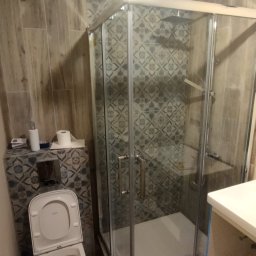 STRONG WALL SP. Z O.O. - Wykończona łazienka z kabiną prysznicową z chromowanymi profilami, toaletą z otwartą klapą i wzorzystymi płytkami ceramicznymi w odcieniach szarości i beżu.