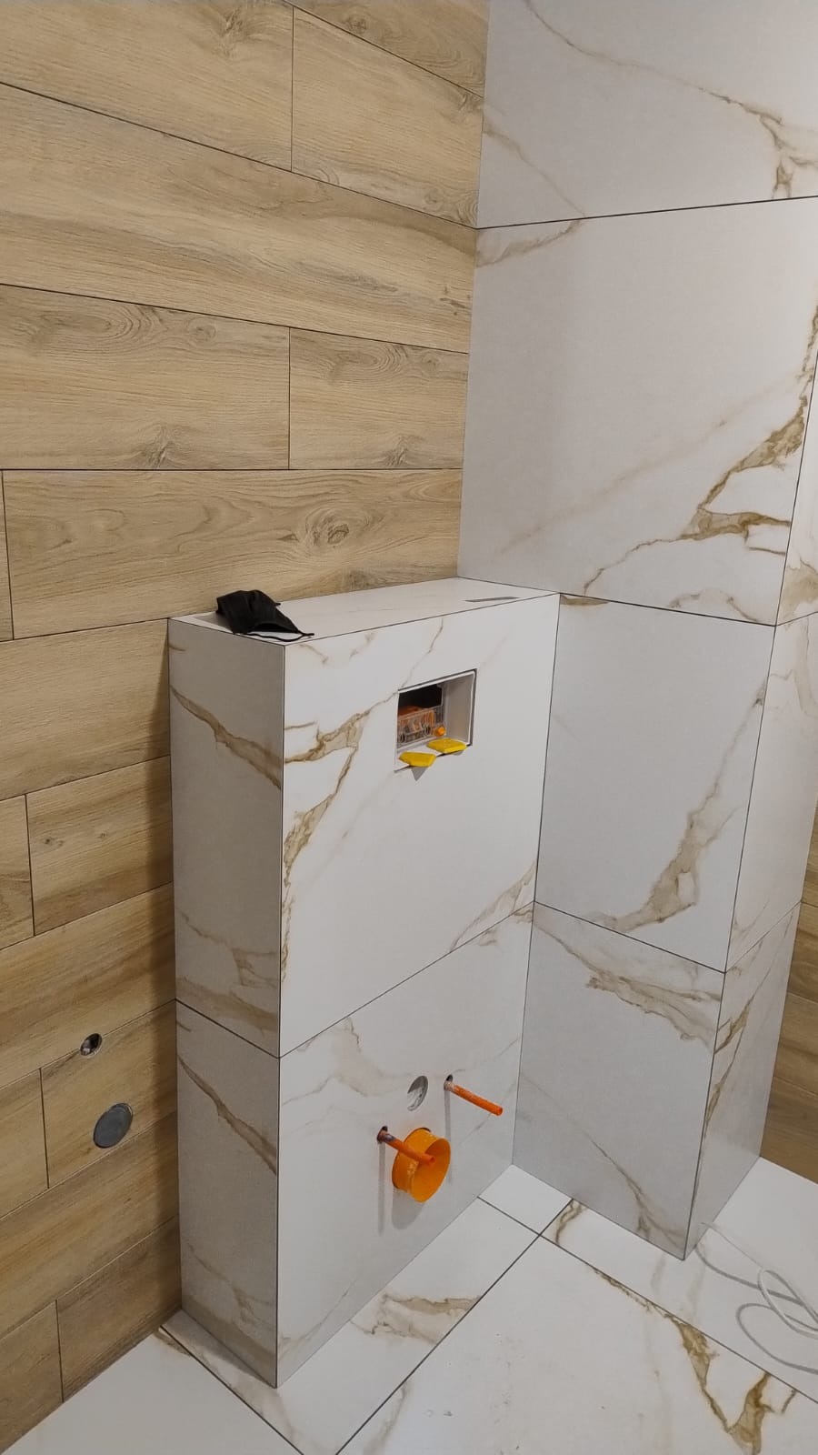 Obudowa stelaża WC w trakcie montażu, wykończona płytkami imitującymi marmur, widoczne elementy instalacji i poziomica, ściana wyłożona panelami drewnopodobnymi.