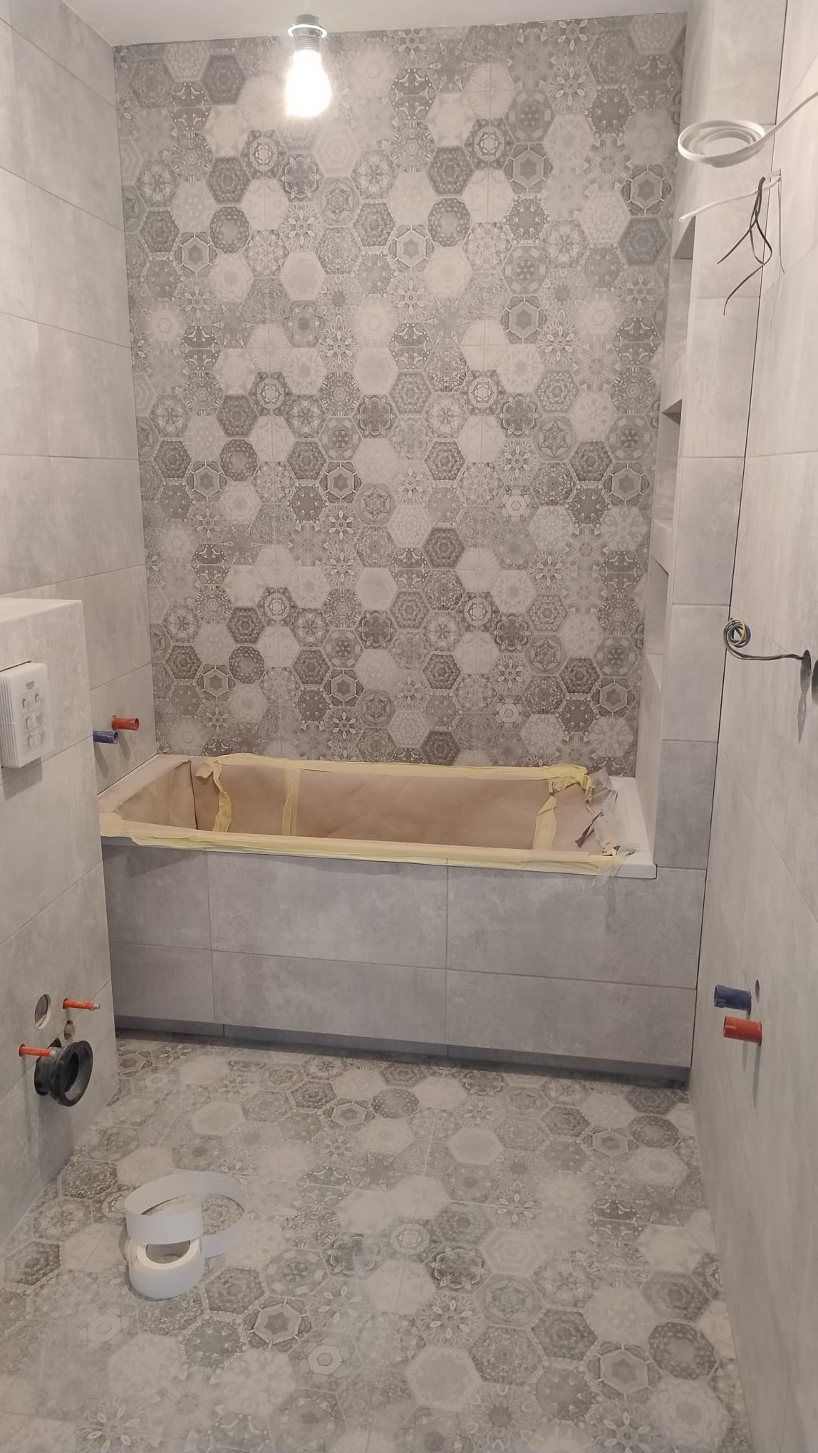 Łazienka w trakcie remontu: szare płytki heksagonalne na podłodze i ścianie, wanna zabezpieczona taśmą malarską, widoczne instalacje hydrauliczne i elektryczne.