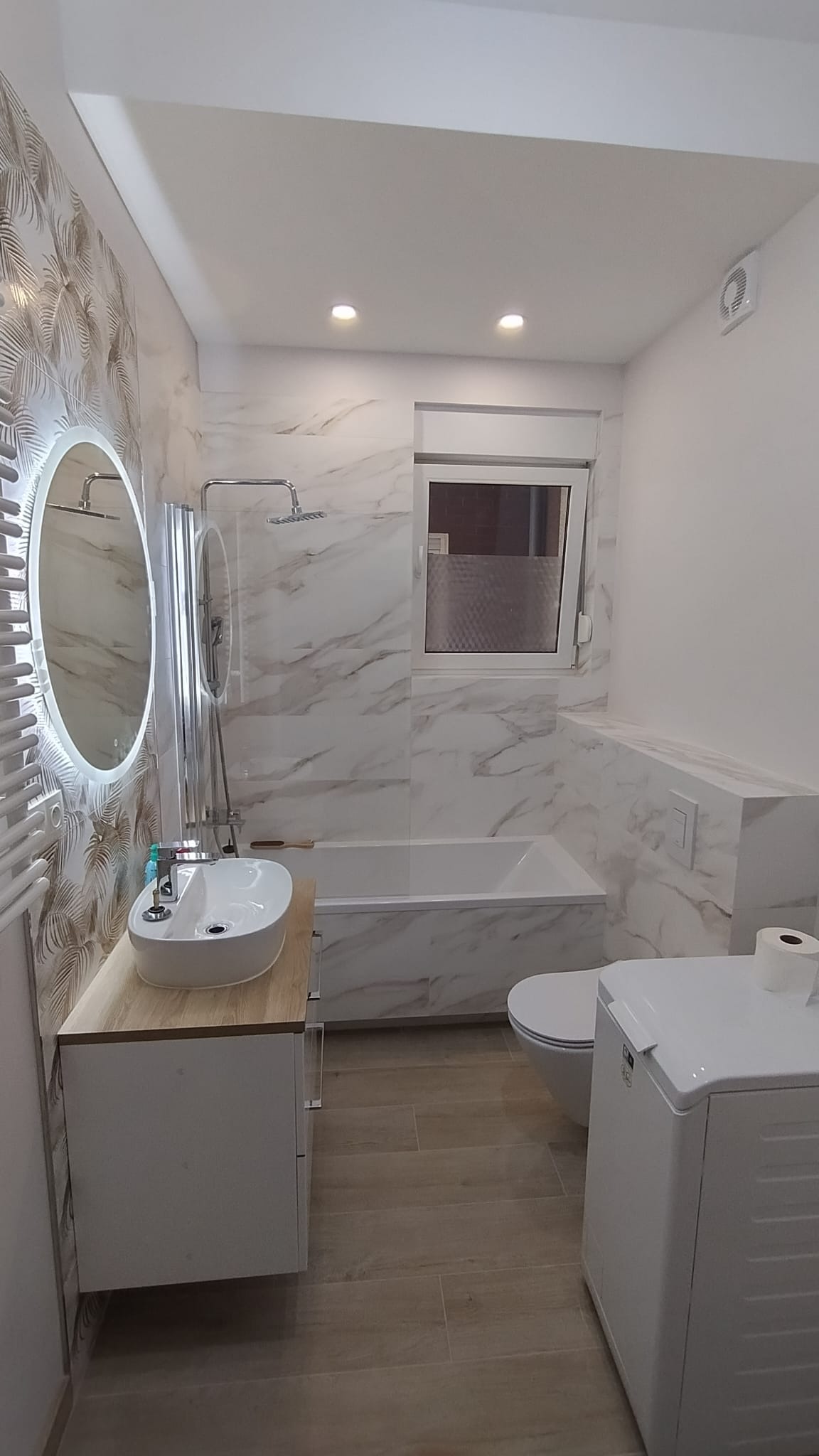 Nowoczesna łazienka po remoncie: marmurowe płytki, okrągłe lustro z podświetleniem LED, drewniany blat umywalki, prysznic za szklaną ścianką, pralka i toaleta.