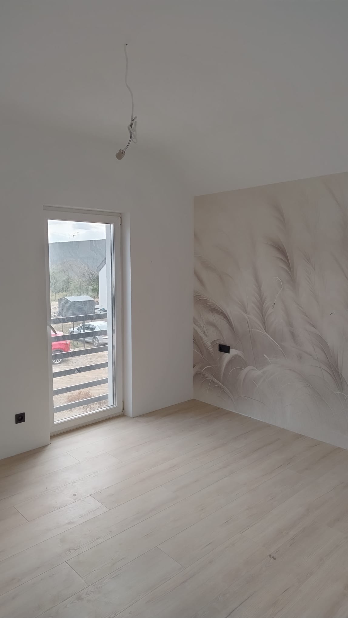 Pusty pokój z drewnianą podłogą, białe ściany, jedna ze ścian z dekoracyjną tapetą w motywie roślinnym, widoczne wystające kable od oświetlenia sufitowego.