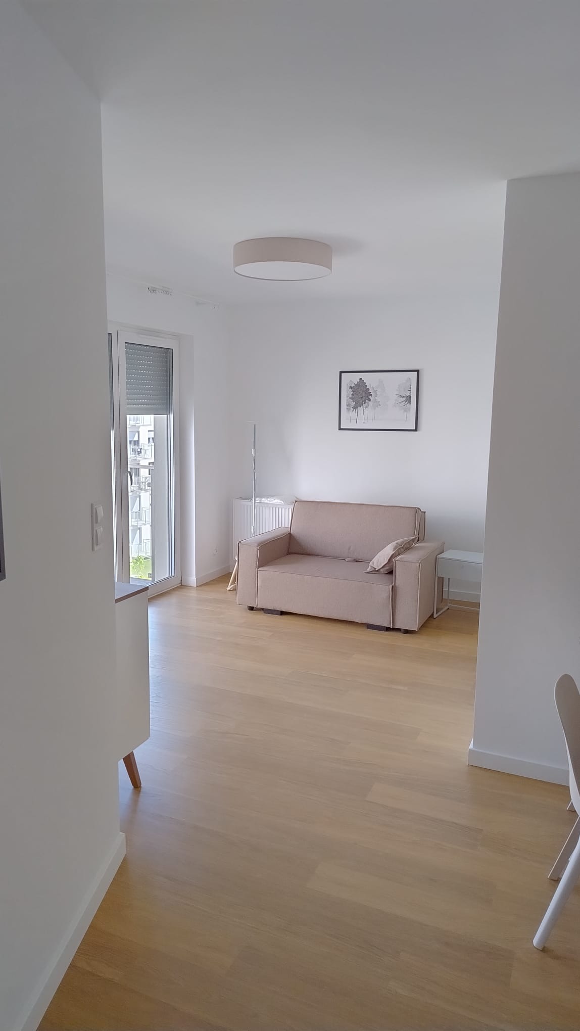 Jasne, minimalistyczne wnętrze po remoncie: beżowa sofa, drewniana podłoga, białe ściany i drzwi balkonowe z roletami.
