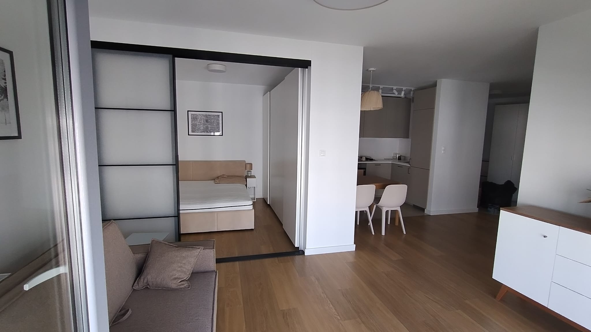 Nowoczesne wnętrze mieszkania z otwartą przestrzenią: sypialnia oddzielona przesuwnymi drzwiami z czarną ramą, widok na kuchnię z jadalnią, drewniana podłoga, minimalistyczny styl.