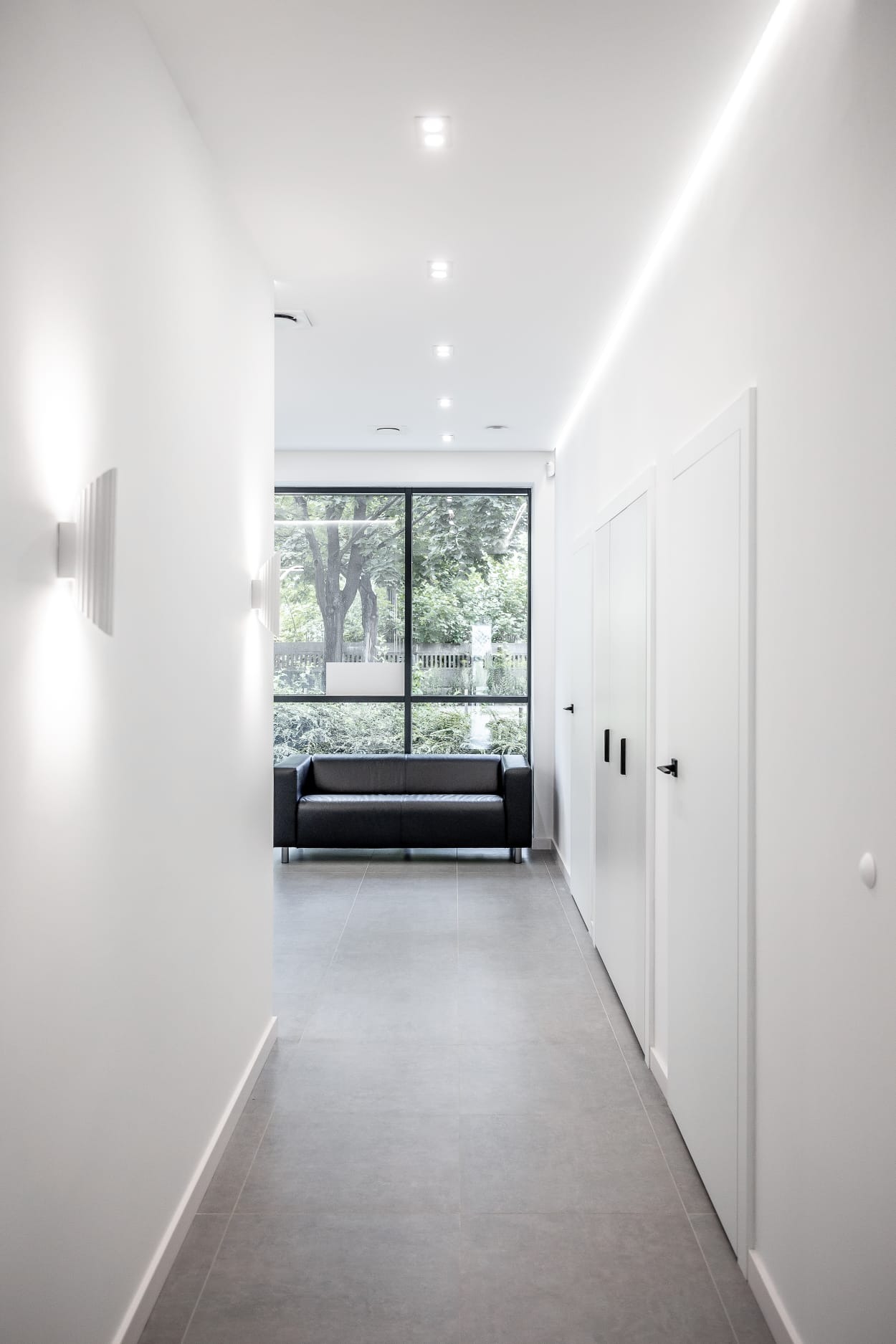 Nowoczesny korytarz z białymi ścianami, szarą podłogą z płytek, czarną sofą pod oknem i minimalistycznym oświetleniem LED na suficie i ścianach.