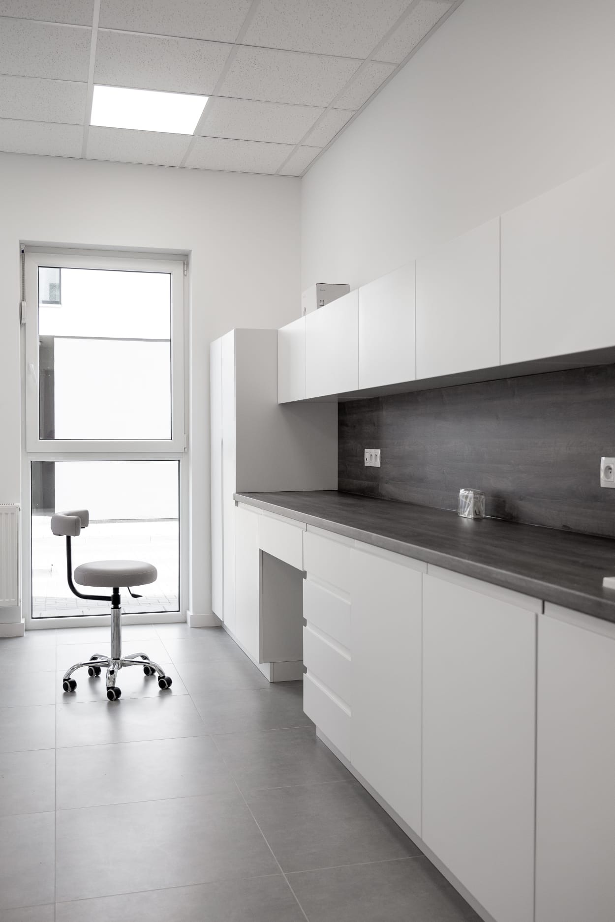 Nowoczesna, minimalistyczna aranżacja wnętrza z białymi szafkami kuchennymi, ciemnym blatem i szarą podłogą z płytek; krzesło na kółkach stoi obok okna.
