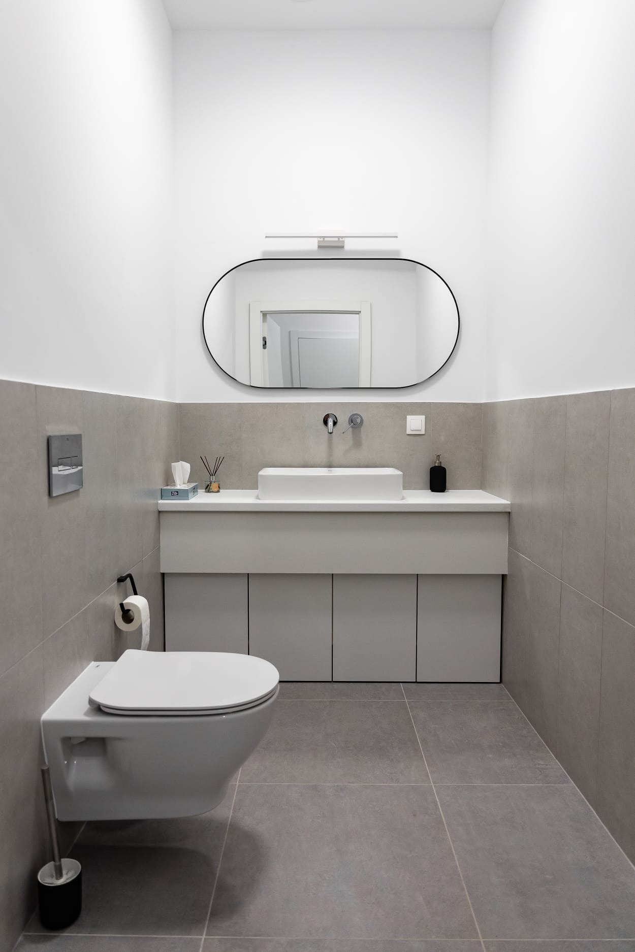 Nowoczesna łazienka w odcieniach szarości i bieli, z podwieszaną toaletą, umywalką nablatową i owalnym lustrem z czarną ramą.