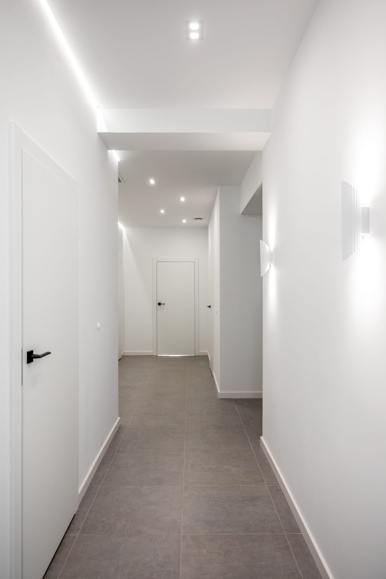 Nowoczesny korytarz z szarą podłogą z płytek, białymi ścianami i drzwiami, oświetlony ledowymi lampami sufitowymi i kinkietami.
