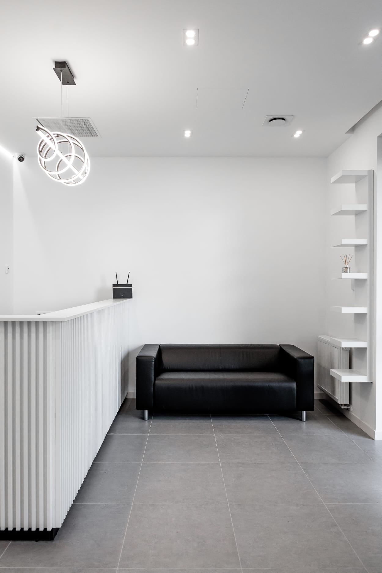 Nowoczesne wnętrze: biała recepcja z dekoracyjnym oświetleniem, czarna skórzana sofa i minimalistyczne białe półki.