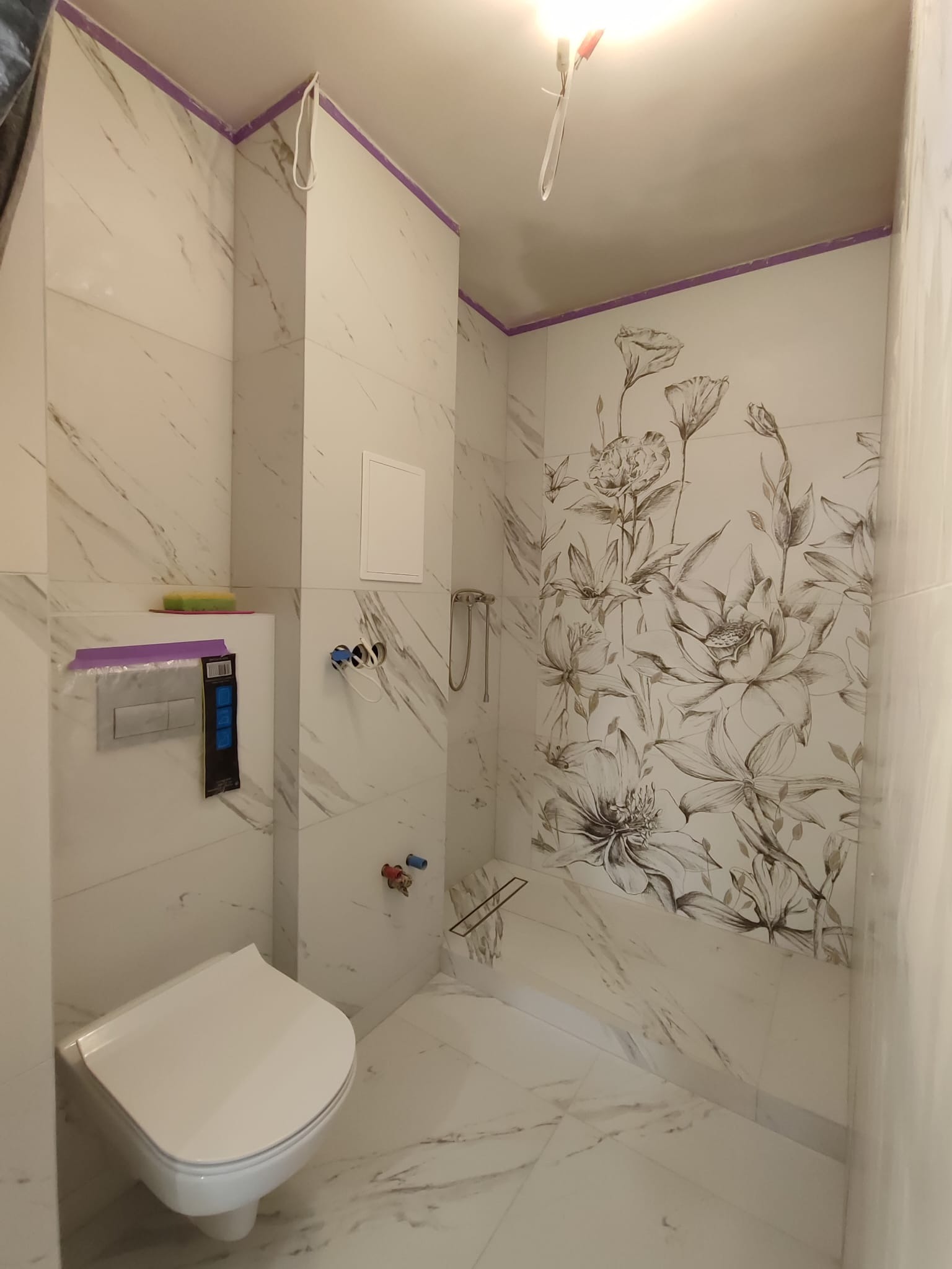 Wnętrze łazienki w trakcie wykończenia, widoczne białe marmurowe płytki z motywem kwiatowym, podwieszana toaleta z zabezpieczeniem, wystające kable i rury, fioletowa taśma malarska na górze ścian.