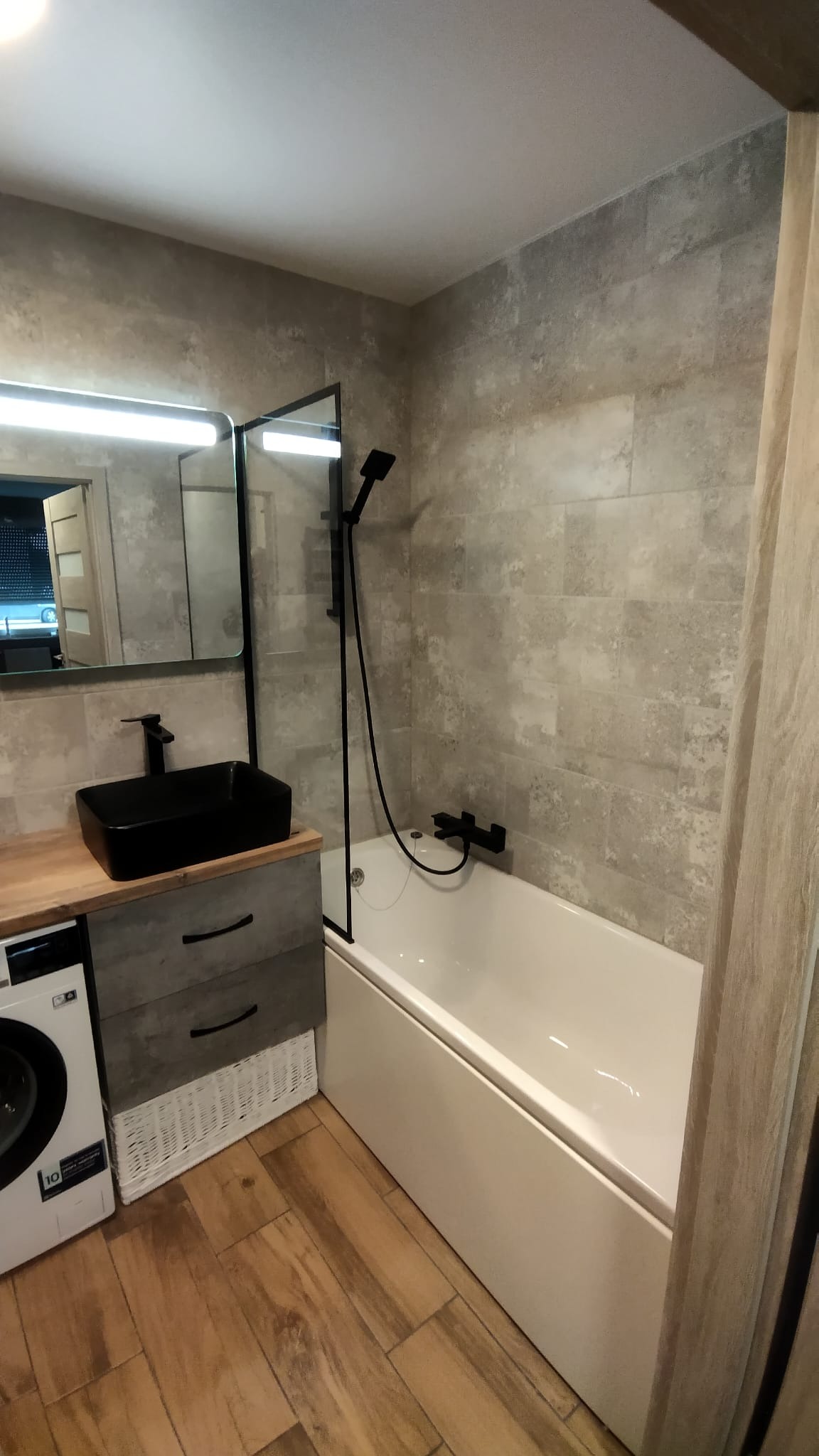 Nowoczesna łazienka z beżowymi płytkami imitującymi kamień, czarną armaturą, prostokątną umywalką nablatową, parawanem nawannowym i drewnopodobną podłogą, pralka w zabudowie.