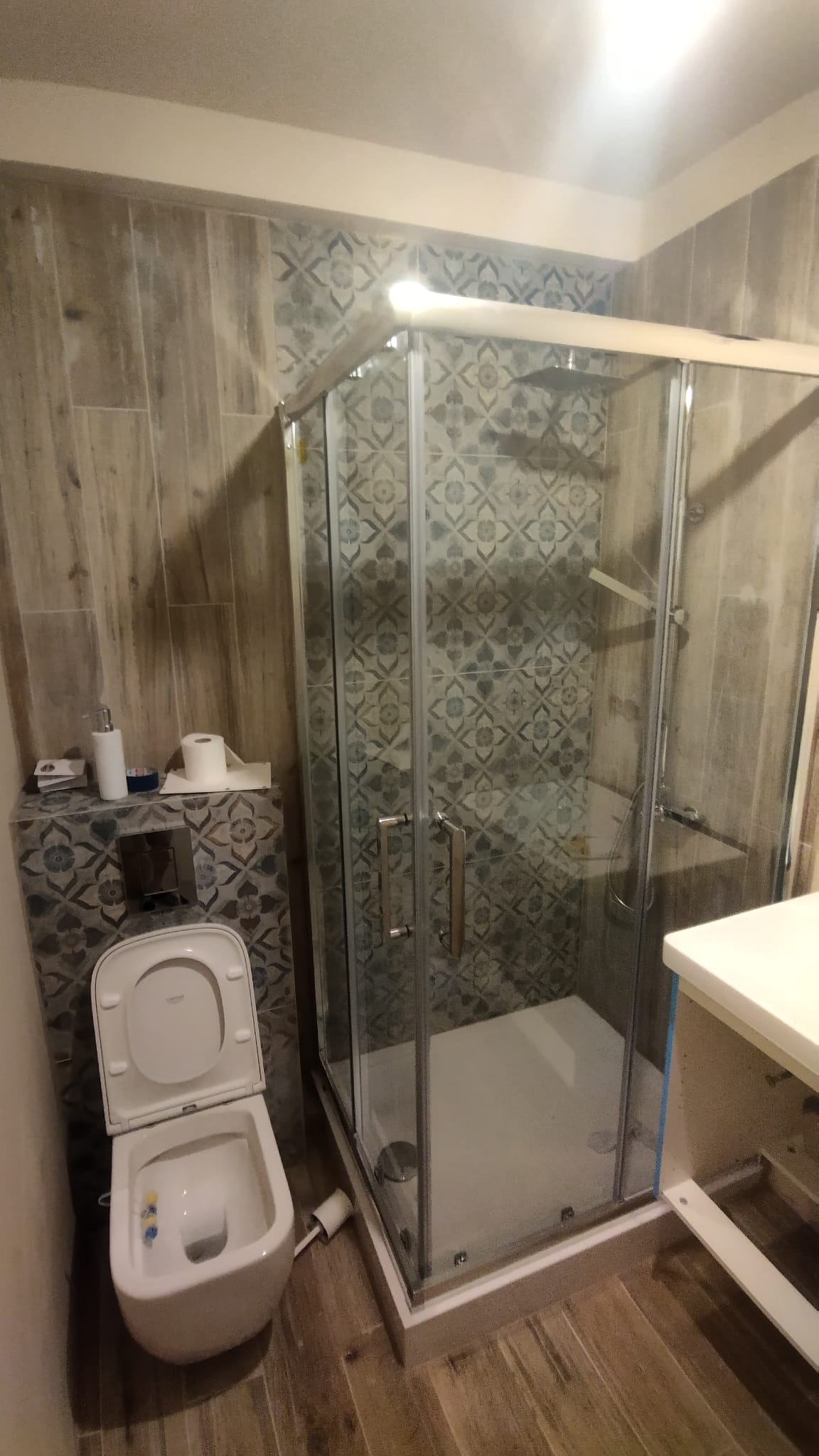 Wykończona łazienka z kabiną prysznicową z chromowanymi profilami, toaletą z otwartą klapą i wzorzystymi płytkami ceramicznymi w odcieniach szarości i beżu.