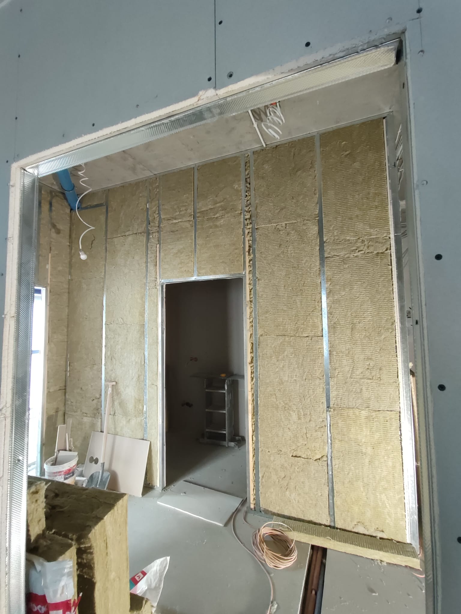 Wnętrze w trakcie prac wykończeniowych: ściany z izolacją z wełny mineralnej, metalowe profile konstrukcyjne, widoczne instalacje elektryczne i hydrauliczne, przygotowanie do montażu płyt...