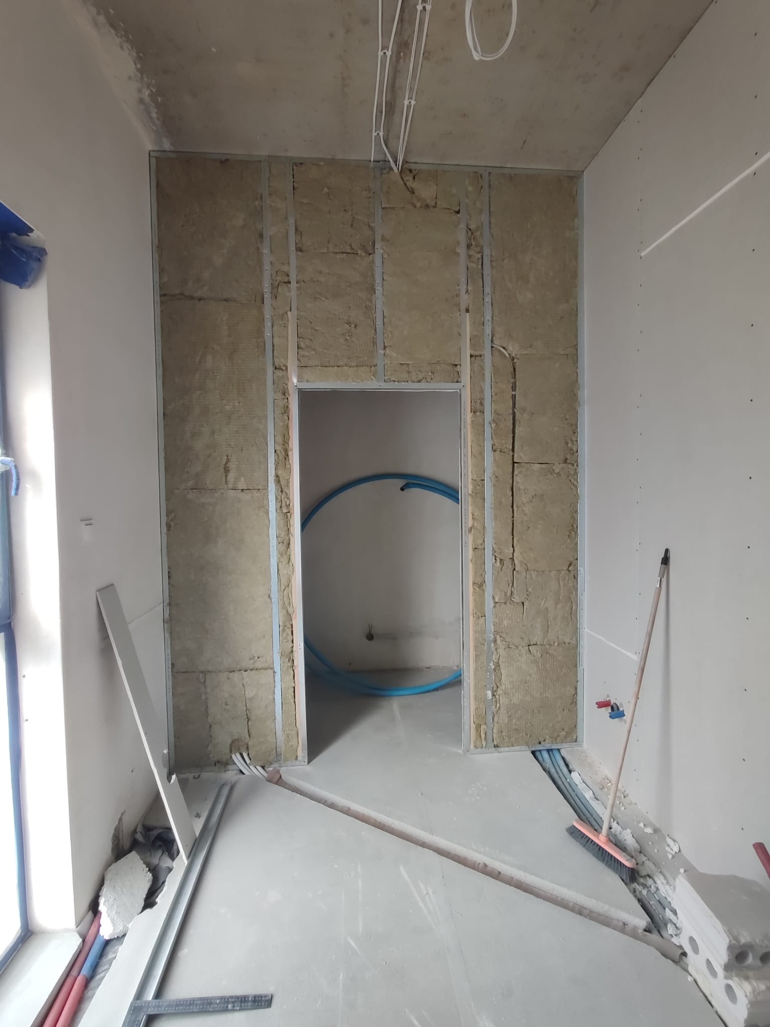 Montaż wełny mineralnej w metalowym stelażu pod przyszłą ścianę z płyt gipsowo-kartonowych, widoczne instalacje w ścianie i rury instalacyjne w przejściu.