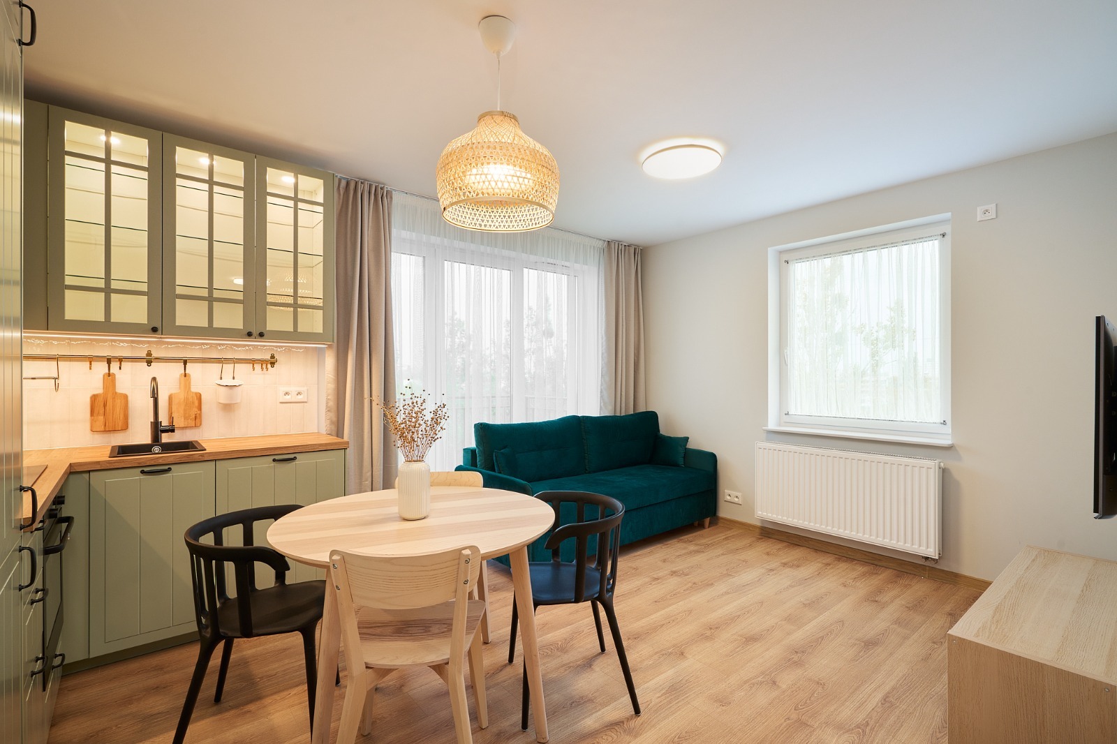 Jasne wnętrze mieszkania z zieloną zabudową kuchenną, okrągłym drewnianym stołem, turkusową sofą i drewnianą podłogą.
