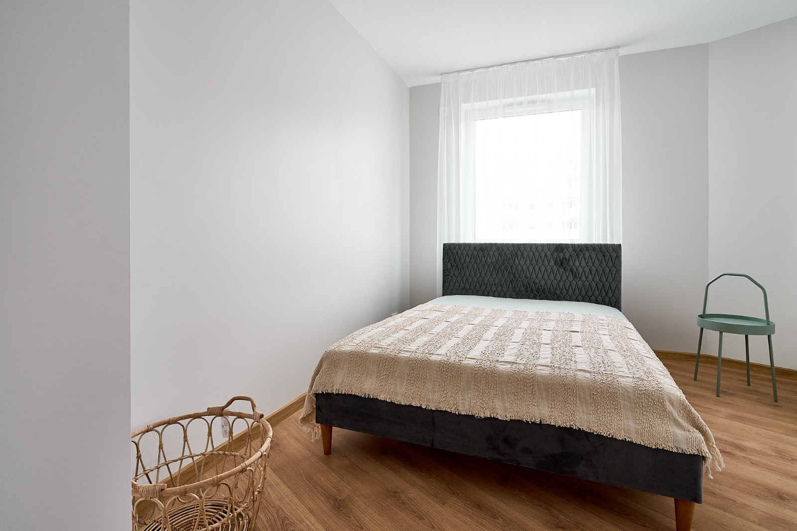 Nowoczesna sypialnia z łóżkiem z ciemnym, pikowanym zagłówkiem, beżową narzutą, drewnianą podłogą i minimalistycznym zielonym krzesłem przy białej ścianie.