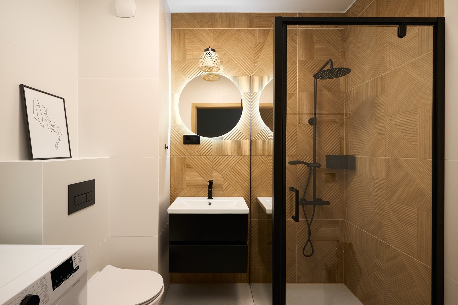 Nowoczesna łazienka z kabiną prysznicową typu walk-in, umywalką na szafce w kolorze czarnym, okrągłym lustrem z podświetleniem LED i dekoracyjnymi płytkami ściennymi w odcieniach brązu, pralka w rogu.