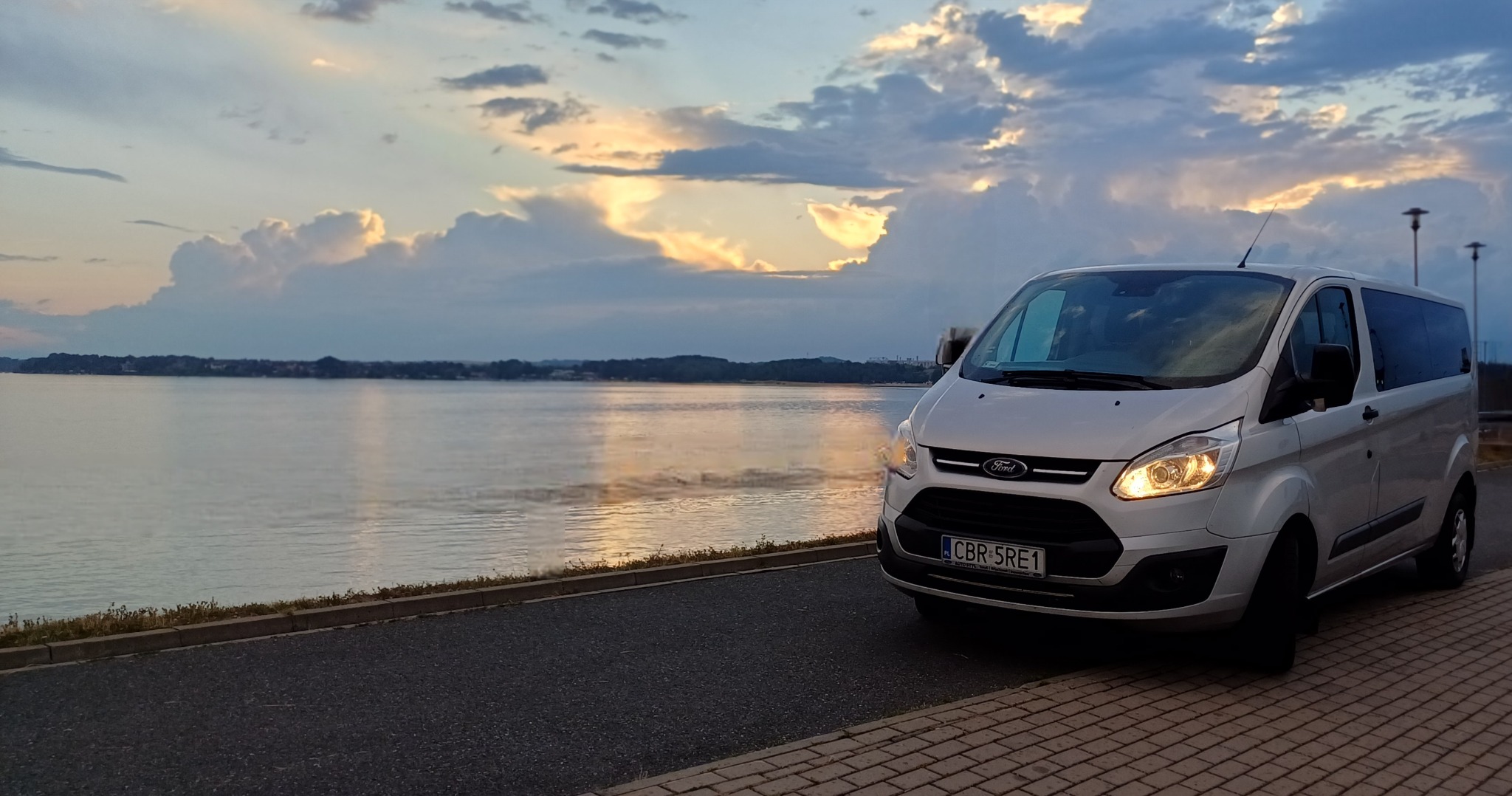 Srebrny bus Ford Transit z włączonymi światłami, zaparkowany nad brzegiem jeziora o zmierzchu, z widocznym zachodzącym słońcem odbijającym się w wodzie i chmurami na niebie.