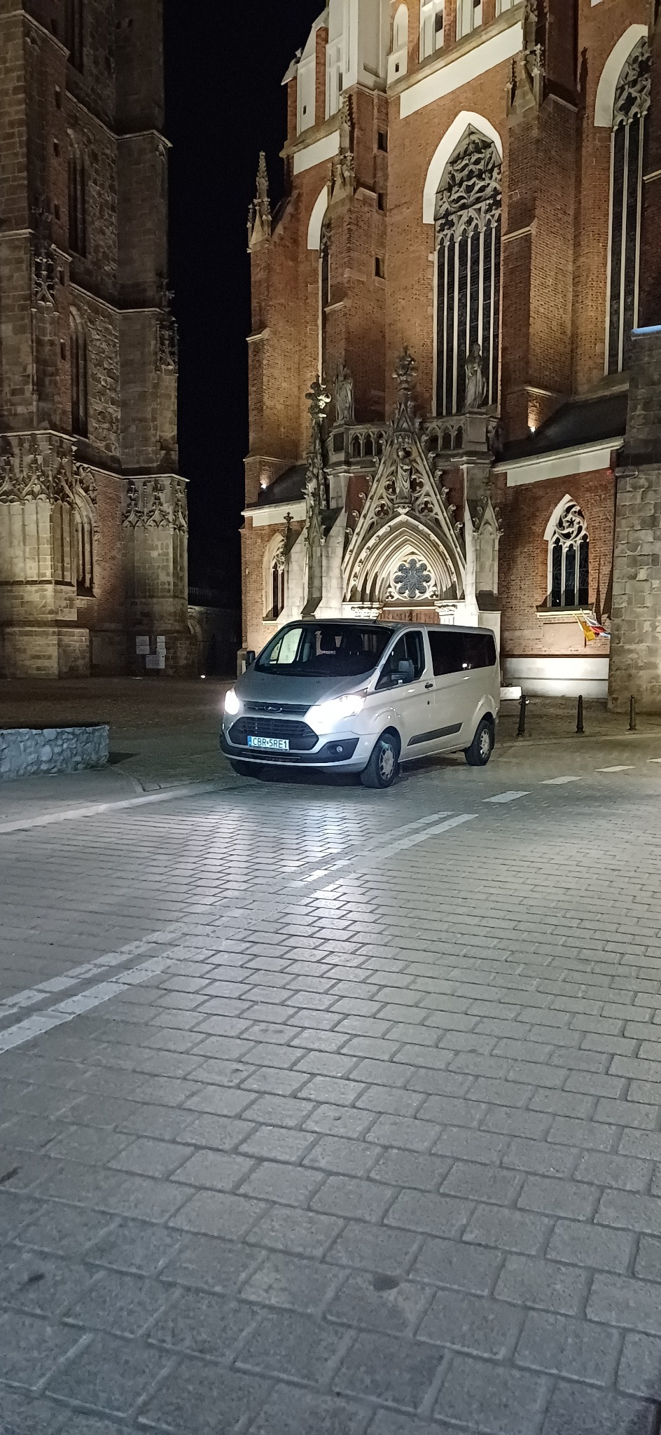 Srebrny van Ford Transit z włączonymi światłami, zaparkowany na brukowanym placu przed ceglanym, gotyckim kościołem nocą.