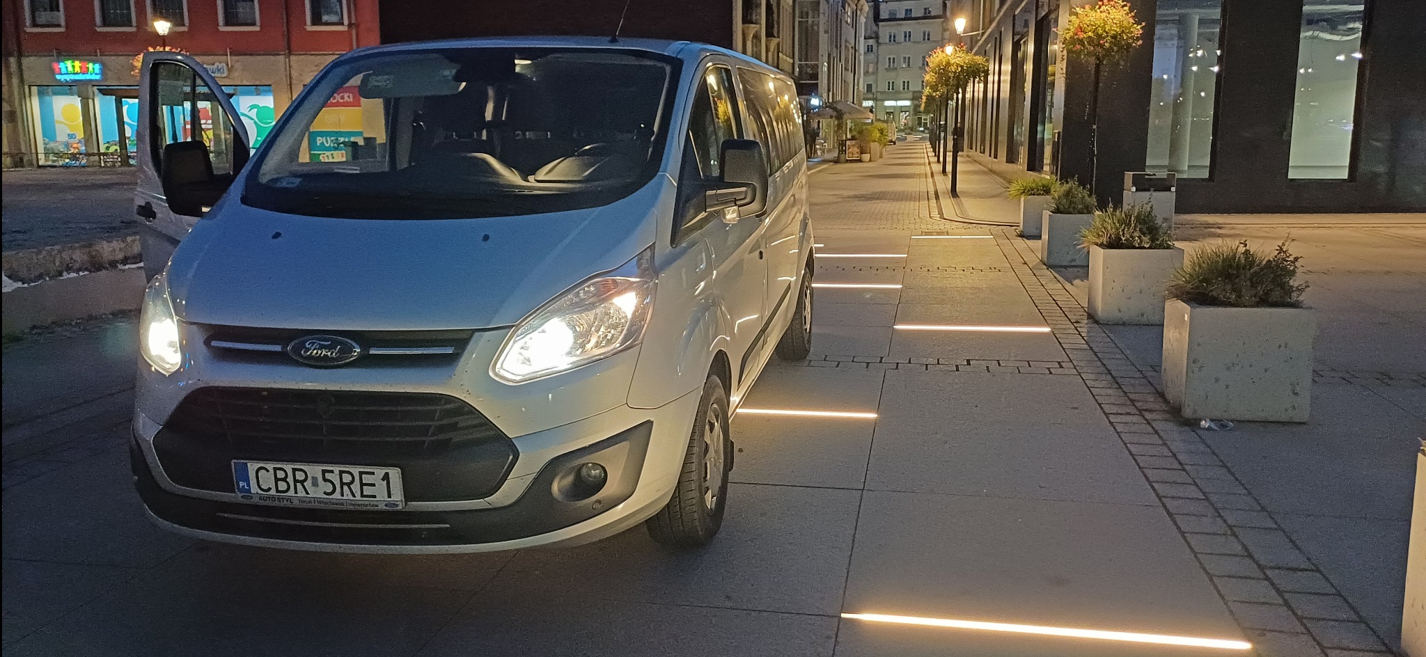 Srebrny Ford Transit z otwartymi drzwiami pasażera, zaparkowany na oświetlonym chodniku w centrum miasta nocą, widoczne donice z roślinami i oświetlenie chodnikowe.