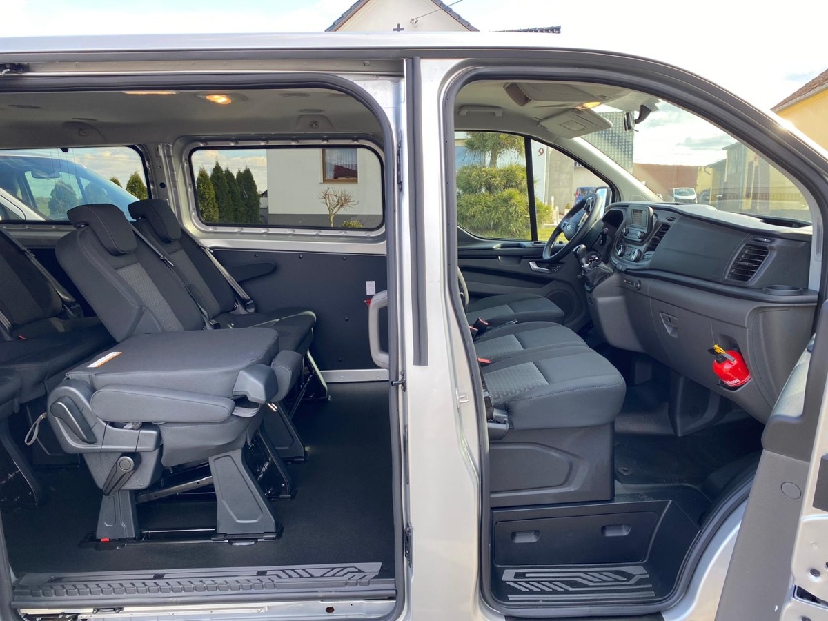 Wnętrze srebrnego busa z rozsuwanymi drzwiami, widoczne fotele pasażerskie z regulacją, deska rozdzielcza i gaśnica.