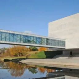 Nowoczesny budynek z betonową elewacją połączony szklanym przejściem nad rzeką, odbicie w wodzie.