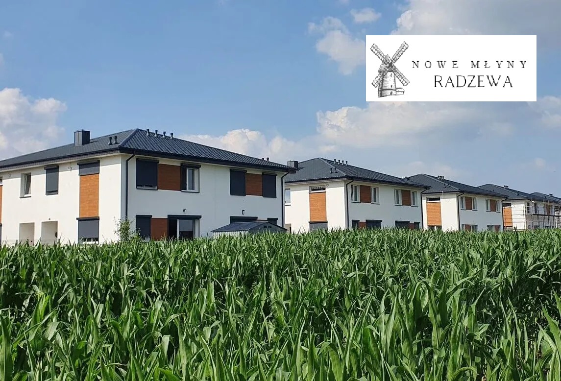 Nowoczesne, dwukondygnacyjne domy szeregowe z ciemnymi dachami i roletami, widoczne zza pola kukurydzy w słoneczny dzień, logo dewelopera w prawym górnym rogu.