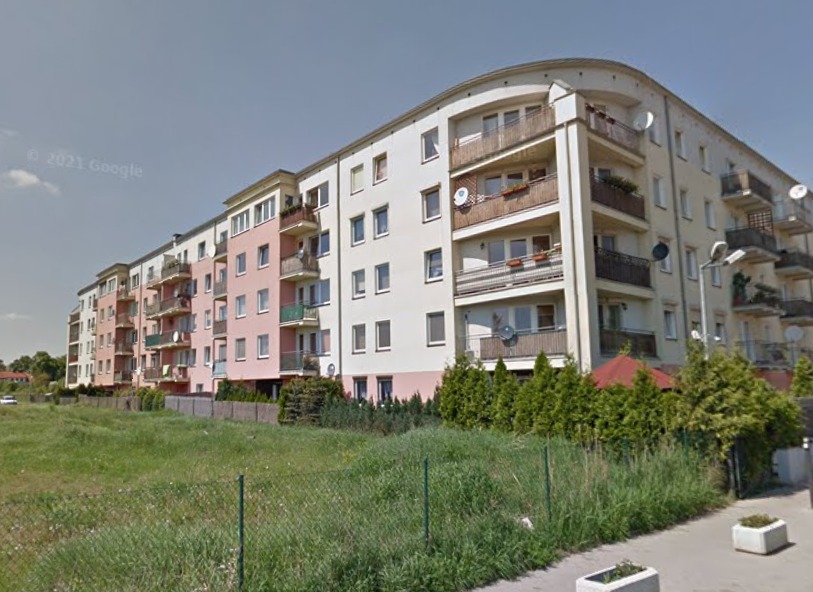 Wielorodzinny budynek mieszkalny z balkonami i antenami satelitarnymi, widok z poziomu ulicy przez metalowe ogrodzenie.