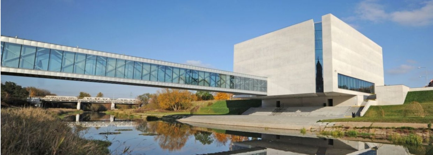 Nowoczesny budynek z betonową elewacją połączony szklanym przejściem nad rzeką, odbicie w wodzie.