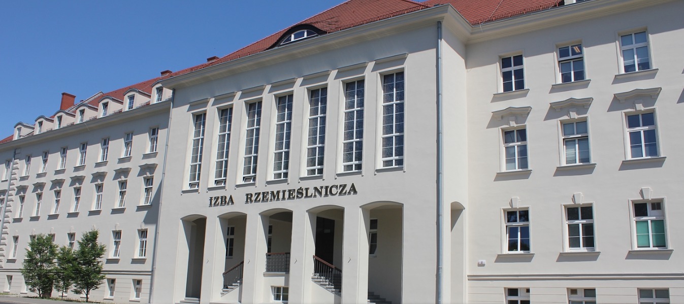 Budynek Izby Rzemieślniczej z czerwonym dachem i licznymi oknami w białych ramach, widoczny z zewnątrz w słoneczny dzień.