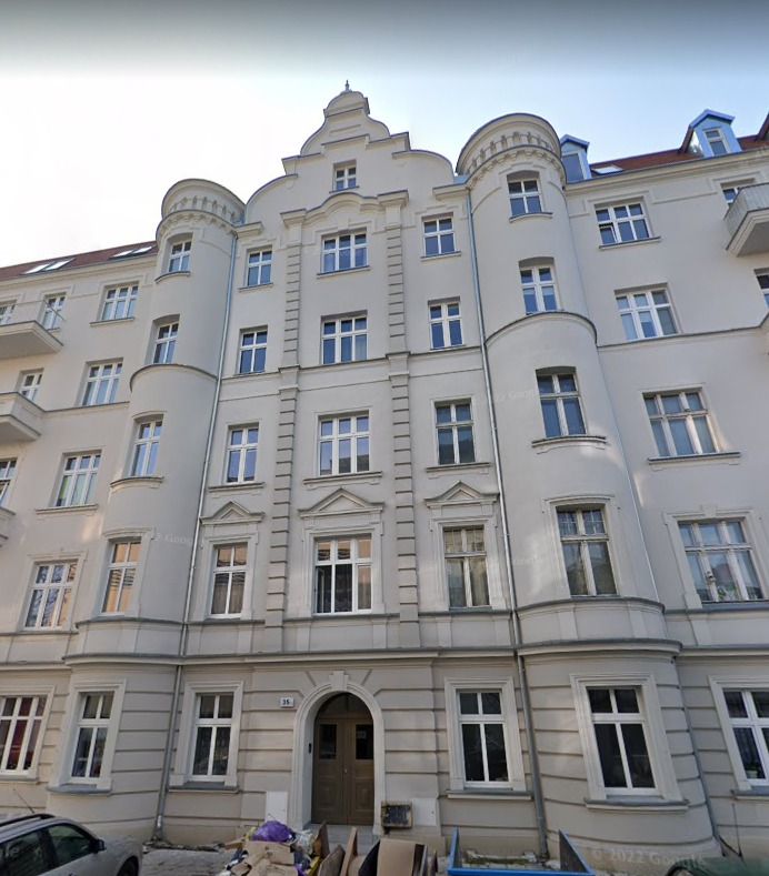 Elewacja kamienicy w stylu secesyjnym z jasną fasadą, widoczne okna z białymi ramami, ozdobne wieżyczki i wejście z numerem 36, przed budynkiem zaparkowany samochód i śmieci.