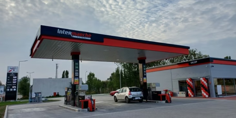 Stacja benzynowa Intermarche z cennikiem paliw, dystrybutorami i zaparkowanym srebrnym samochodem osobowym. W tle budynek sklepu z dekoracją balonową.