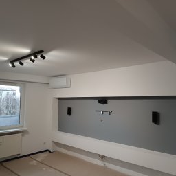 Piotr Wiktorowski - Szare wnętrze pokoju z oknem, grzejnikiem i przygotowaną ścianą pod sprzęt audio-video, oświetlenie sufitowe. Widoczne zabezpieczenia podłogi.